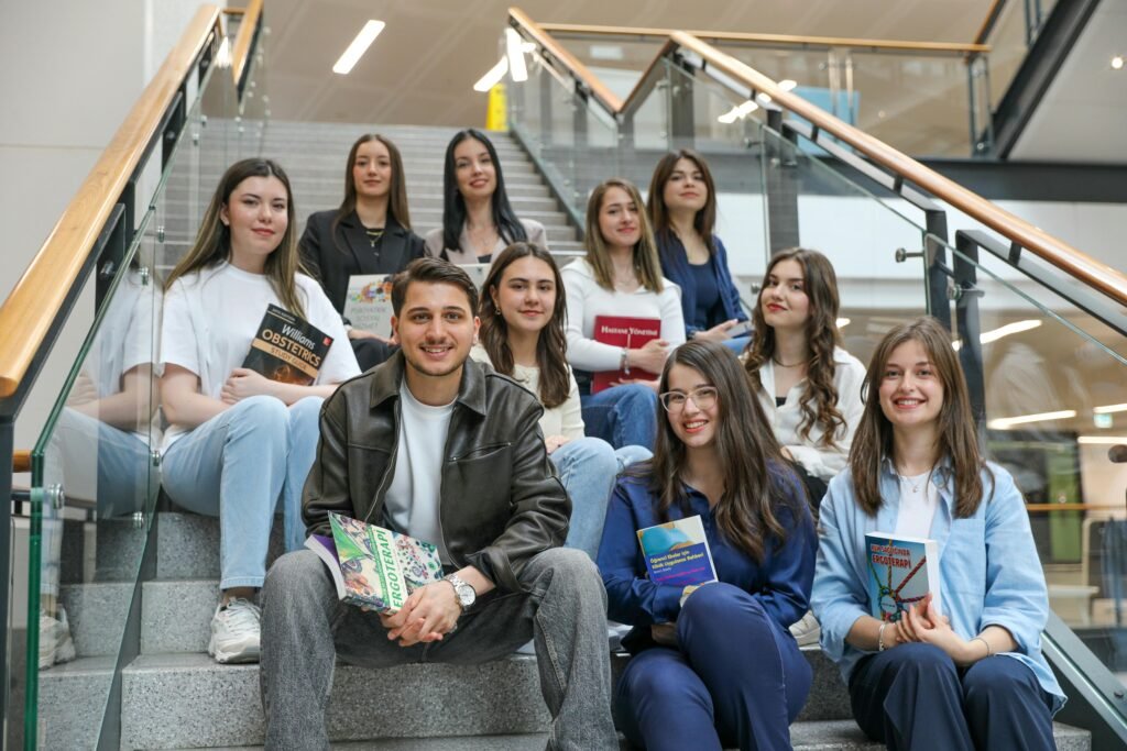 A diverse group of students sitting on stairs indoors, holding books, and smiling.